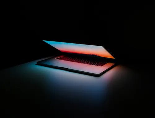 Photo of Mac laptop, partially closed, with screen aglow in a dark room.
