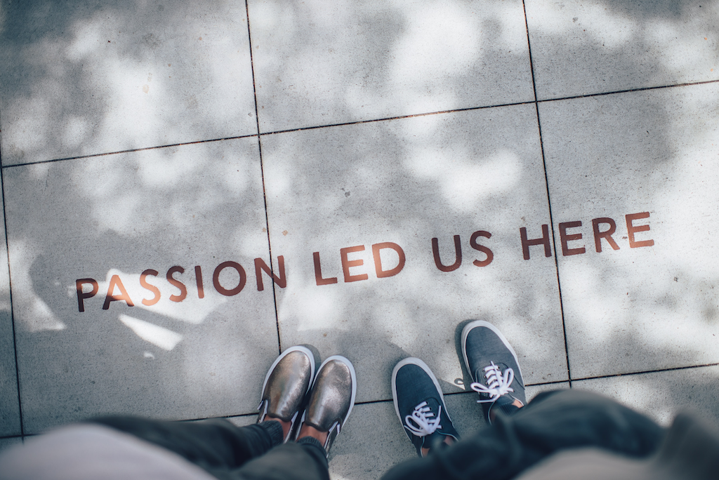 Photo of the feet of two people, standing on a sidewalk that has the words, "PASSION LED US HERE" engraved in it. 