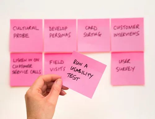 A hand holding a sticky note with "Run a usability test" in front of a wall with sticky notes.