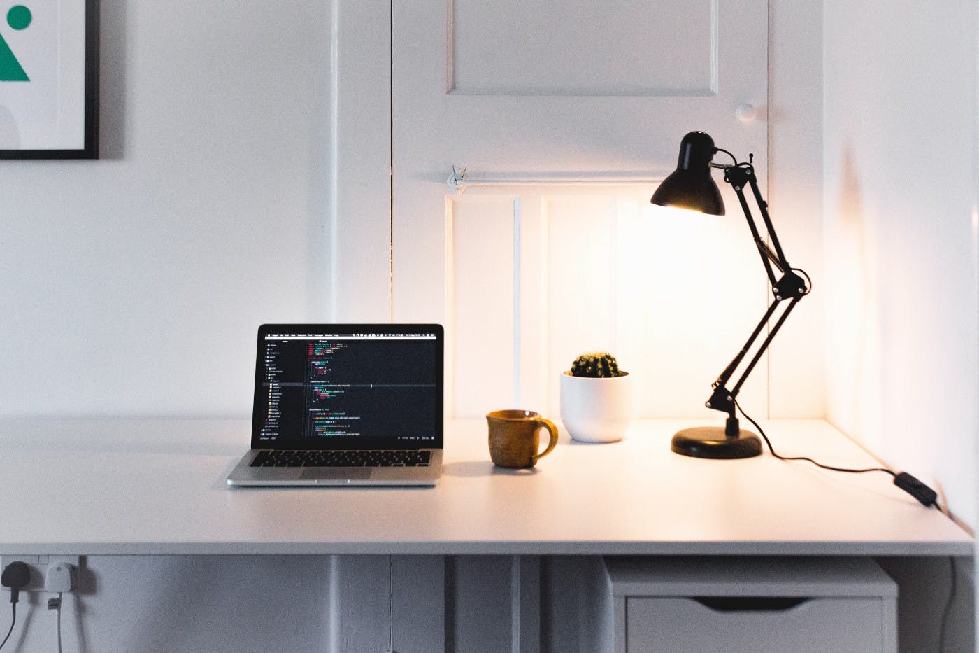 A white desk with an Apple laptop, coffee mug and a lamp.